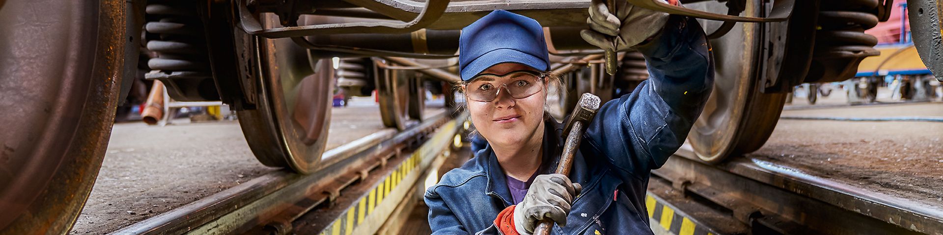 Eine Industriemechanikerin in Arbeitskleidung steht in der Wartungsgrube unter einem Zug und hält einen Hammer