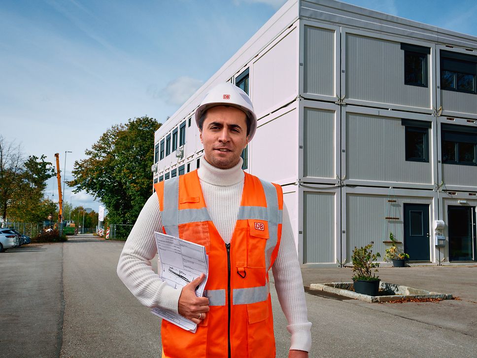 Ein Bauüberwacher in Warnkleidung und mit Helm hat Unterlagen in der Hand und läuft an einem Baucontainer vorbei