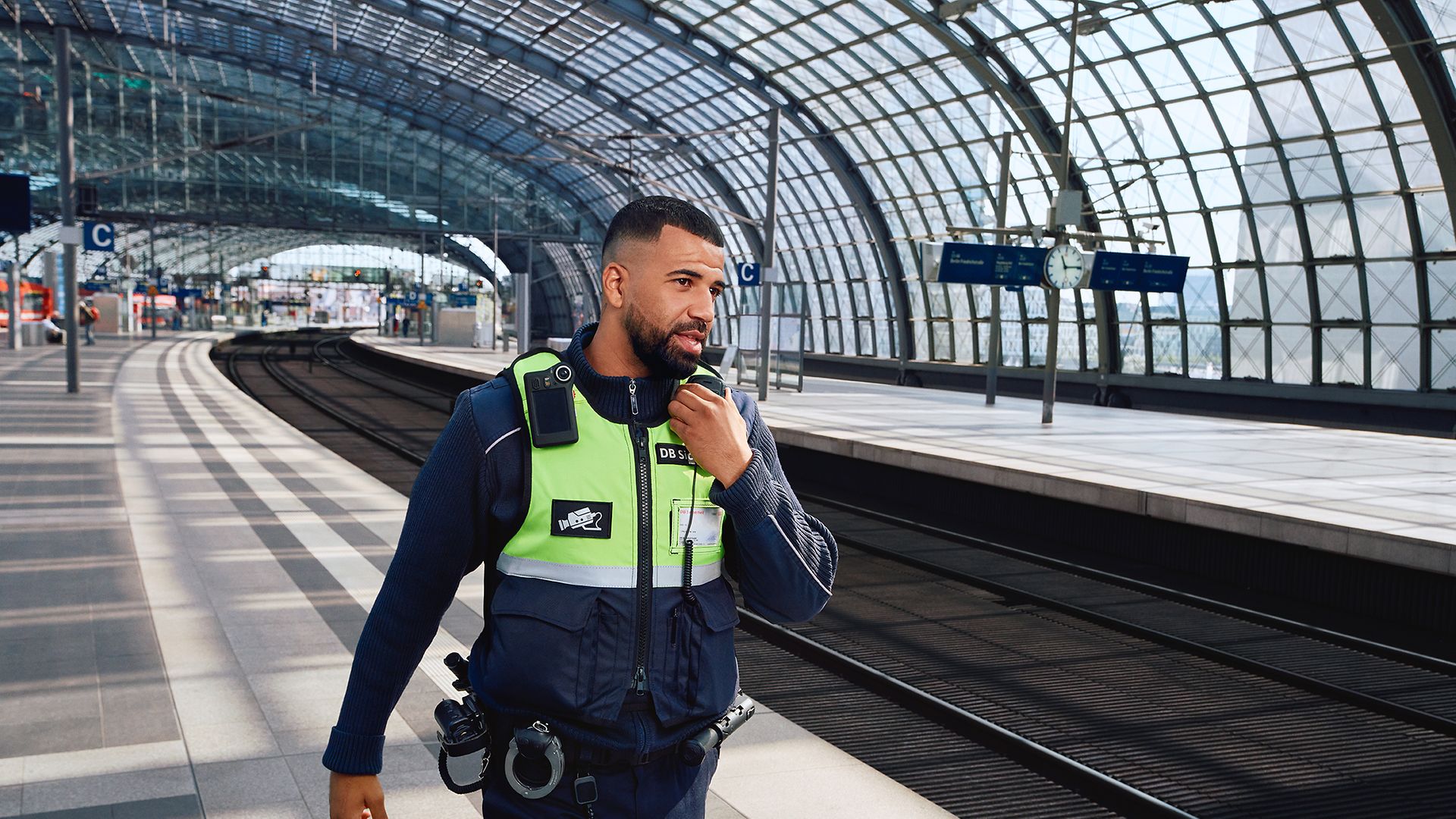 Eine Fachkraft für Schutz und Sicherheit läuft am Bahnsteig eines Bahnhofs entlang und spricht in sein Funkgerät