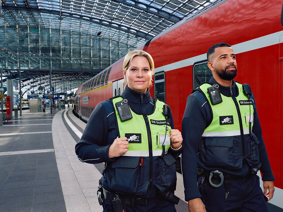 Zwei Fachkräfte für Schutz und Sicherheit laufen am Bahnsteig eines Bahnhofs an einem Regionalzug vorbei