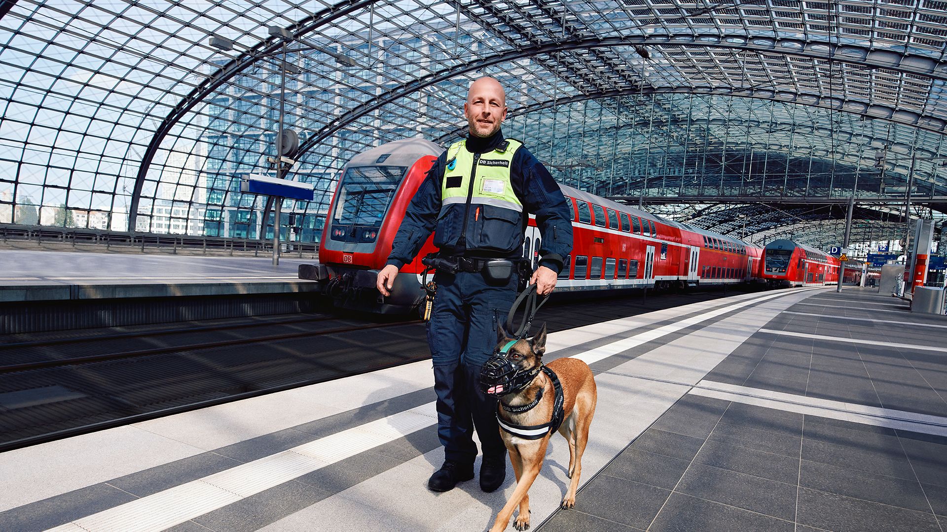 Ein Hundeführer in Unternehmensbekleidung läuft mit einem Wachhund am Bahnsteig eines Bahnhofs an einem Regionalzug vorbei