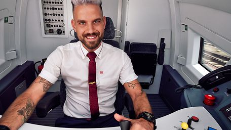 Ein Lokführer im Fernverkehr in Unternehmensbekleidung sitzt im Führerstand eines ICE