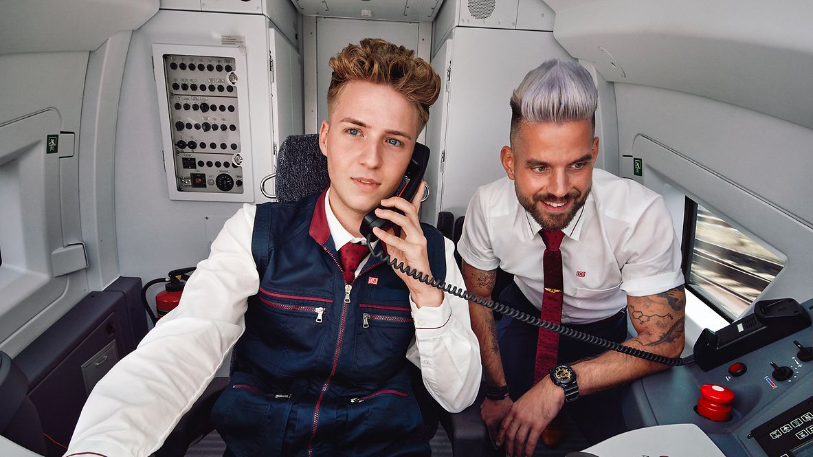 Ein Auszubildender zum Lokführer sitzt neben einem Ausbilder im Führerstand eines ICE und telefoniert über den Zugfunk