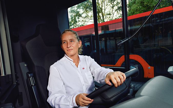 Eine Busfahrerin in Unternehmensbekleidung sitzt am Steuer des Busses