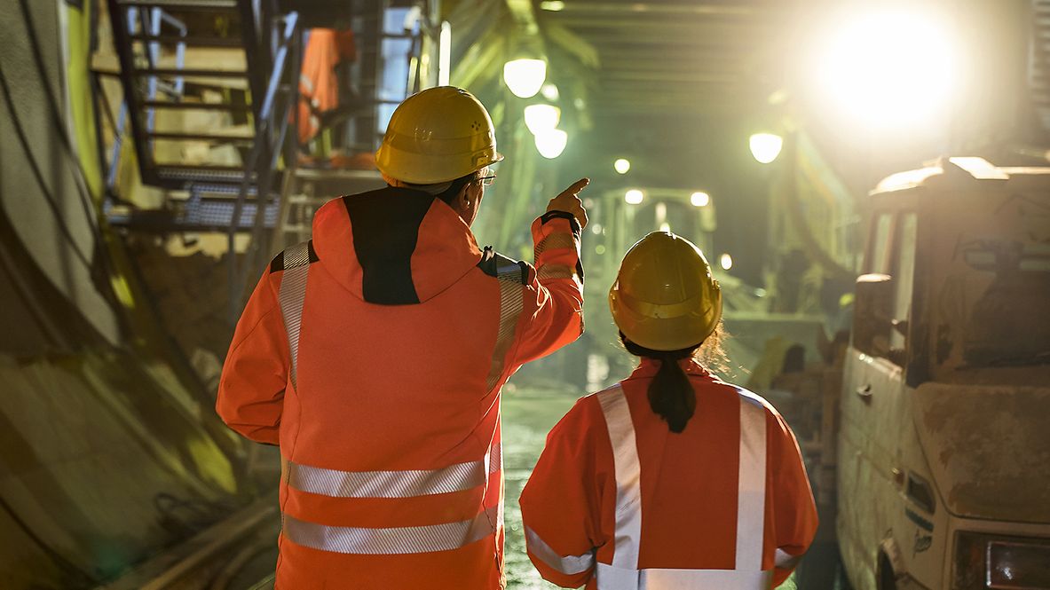 Zwei Ingenieure stehen in Warnkleidung und mit Schutzhelm zeigen auf eine Baustelle