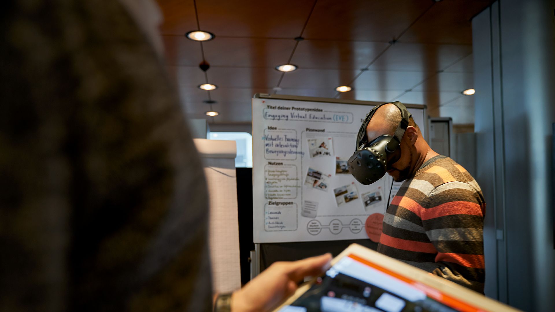 Ein Mann steht in einem Besprechungsraum und trägt eine VR-Brille während eine Frau mit einem Tablet vor ihm steht