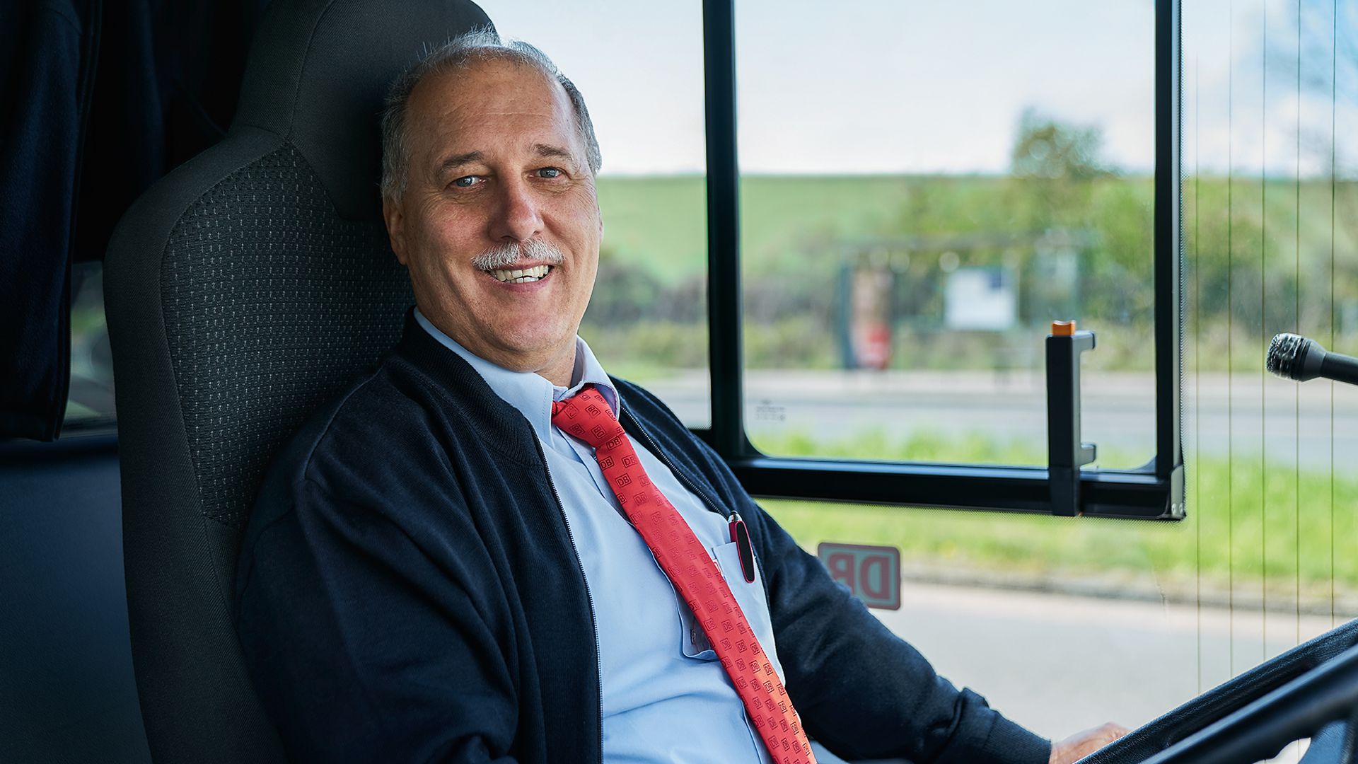 Ein Busfahrer sitzt im Fahrersitz seines Busses und lacht.