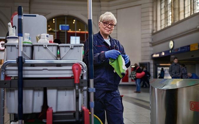 Eine Gebäudereinigerin steht im Bahnhofsgebäude neben einem Reinigungswagen und hält Reinigungsmaterial in der Hand
