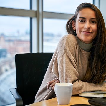 Eine Frau sitzt in einem Büro, mit Ausblick auf Gleise, vor einem Laptop und lächelt.