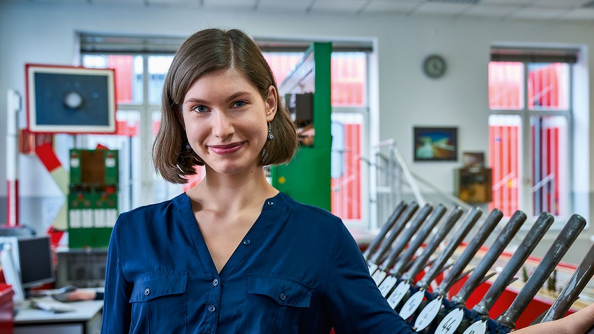 Eine Auszubildende zur Fahrdienstleiterin steht im mechanischen Stellwerk und hat eine Hand an einen Stellwerkshebel gelegt