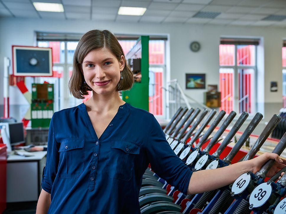 Eine Auszubildende zur Fahrdienstleiterin steht im mechanischen Stellwerk und hat eine Hand an einen Stellwerkshebel gelegt