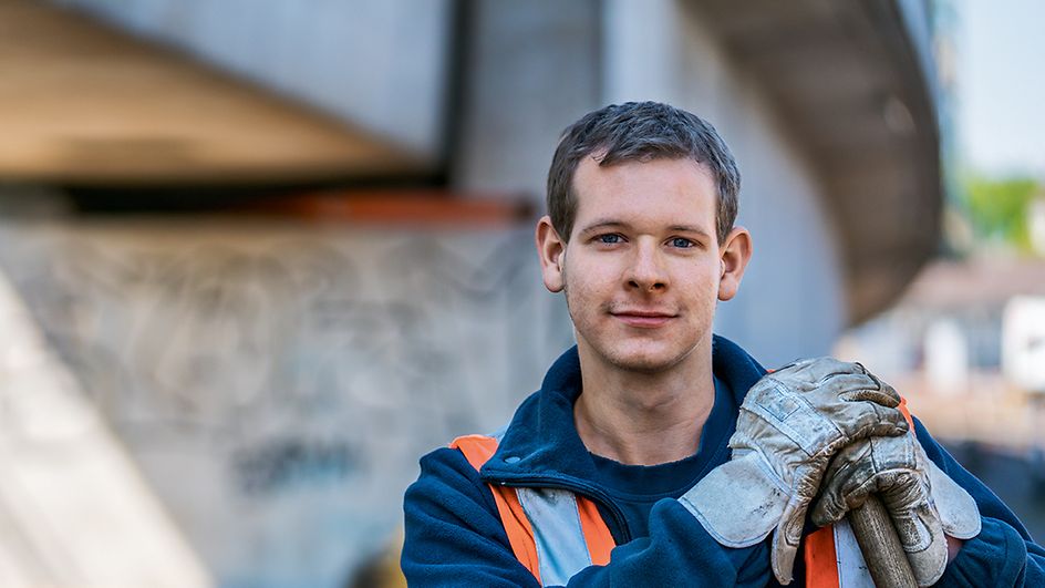 Ein Auszubildender zum Gleisbauer steht in Arbeitskleidung auf ein Gleisbaugerät gestützt vor einer Brücke