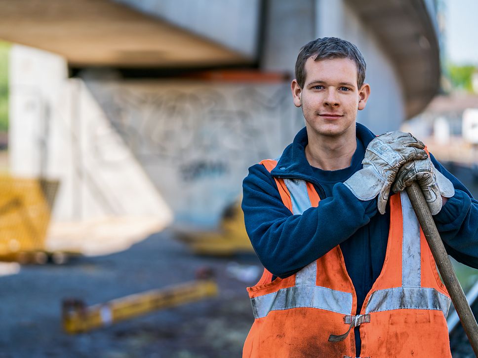 Ein Auszubildender zum Gleisbauer steht in Arbeitskleidung auf ein Gleisbaugerät gestützt vor einer Brücke