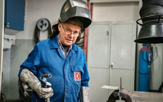 Ein Industriemechaniker steht in der Werkstatt, trägt eine Schweißermaske und hält ein Schweißgerät in der Hand