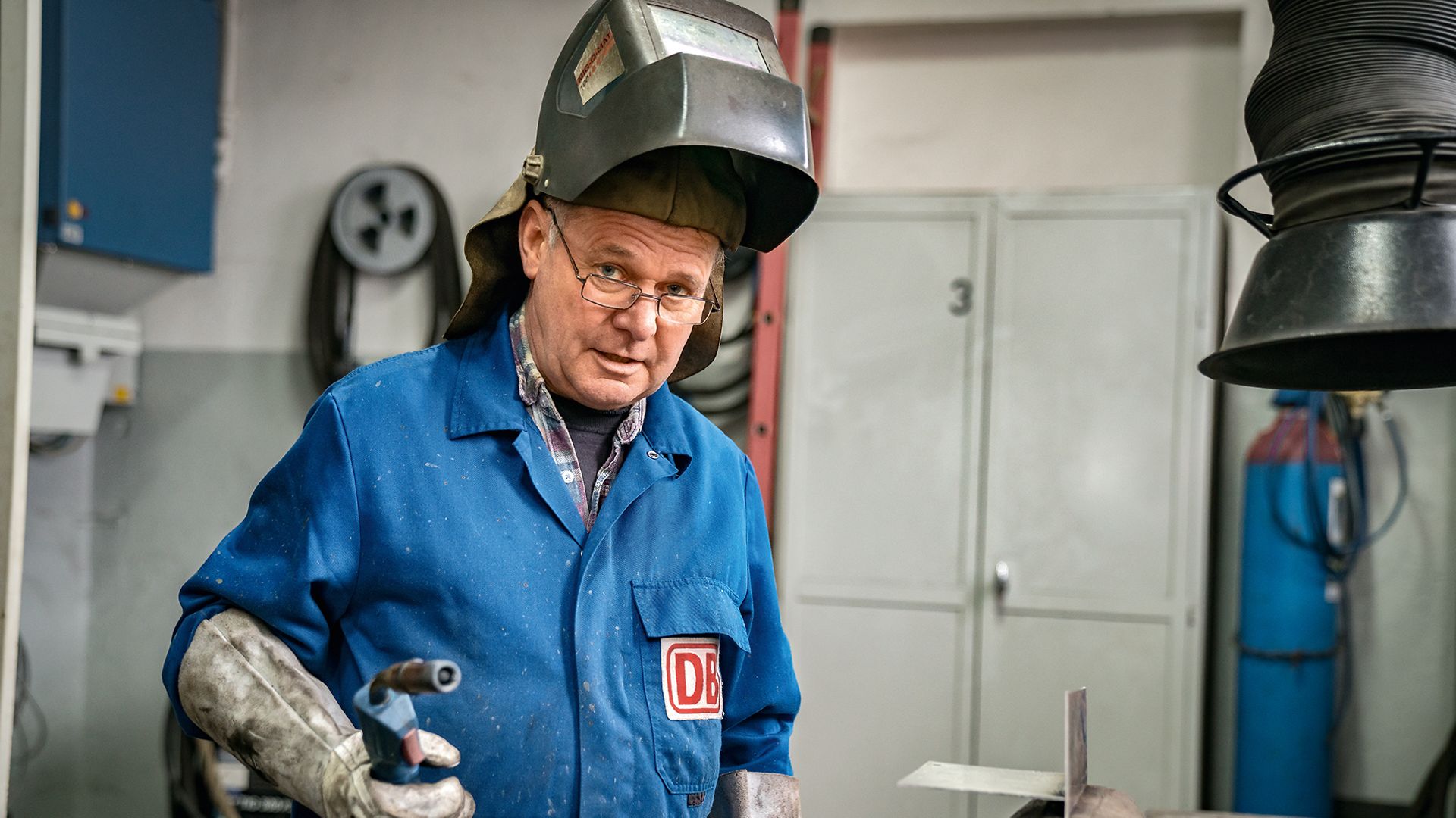 Ein Industriemechaniker steht in der Werkstatt, trägt eine Schweißermaske und hält ein Schweißgerät in der Hand