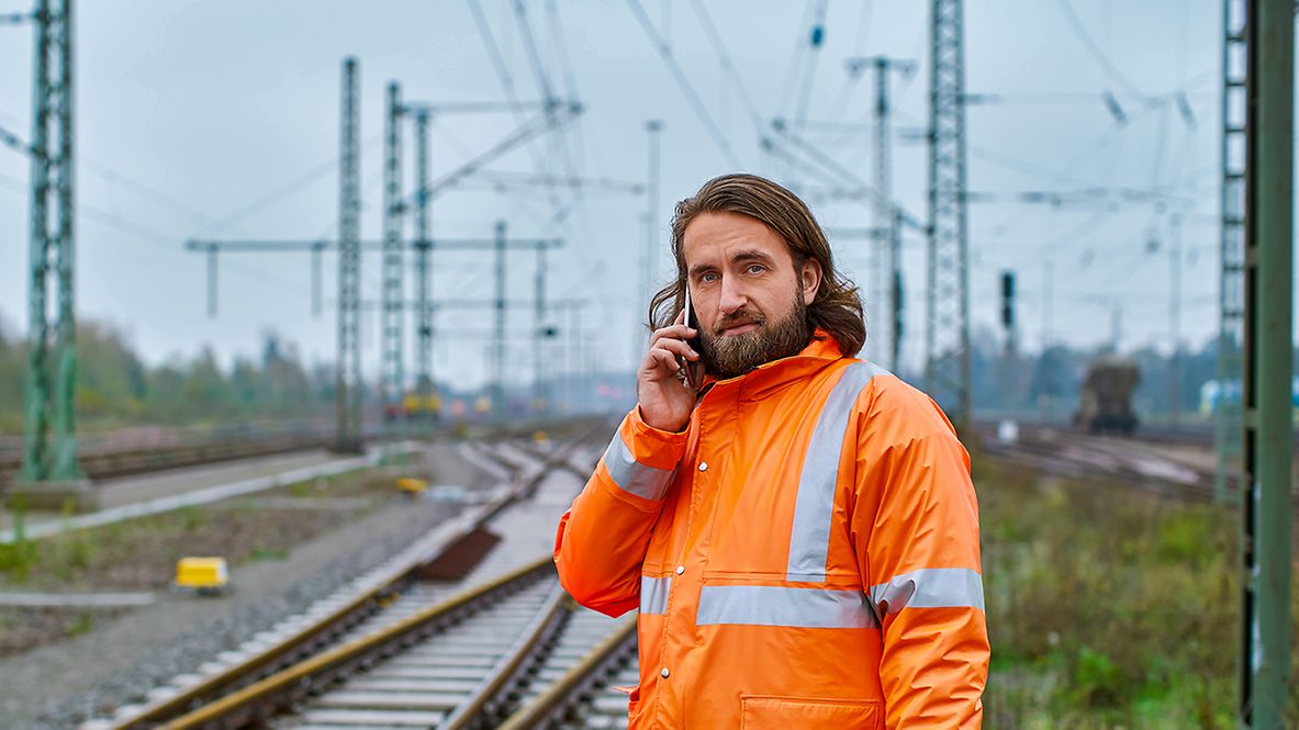 Ein Elektroingenieur steht in Warnkleidung am Gleis und telefoniert