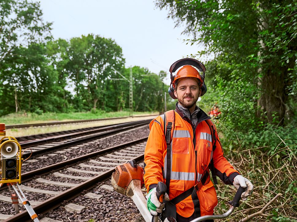 Ein Vegetationspfleger in Warnkleidung steht neben einem Gleis am Waldrand und hält eine Motorsäge in der Hand