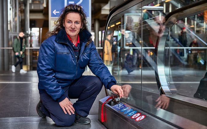 Eine Servicetechnikerin in Arbeitsbekleidung kniet am Bahnhof neben einer Rolltreppe