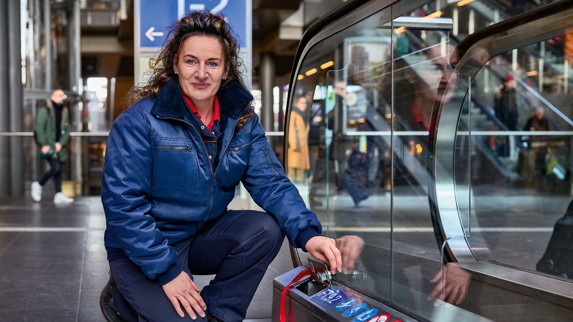 Eine Servicetechnikerin in Arbeitsbekleidung kniet am Bahnhof neben einer Rolltreppe