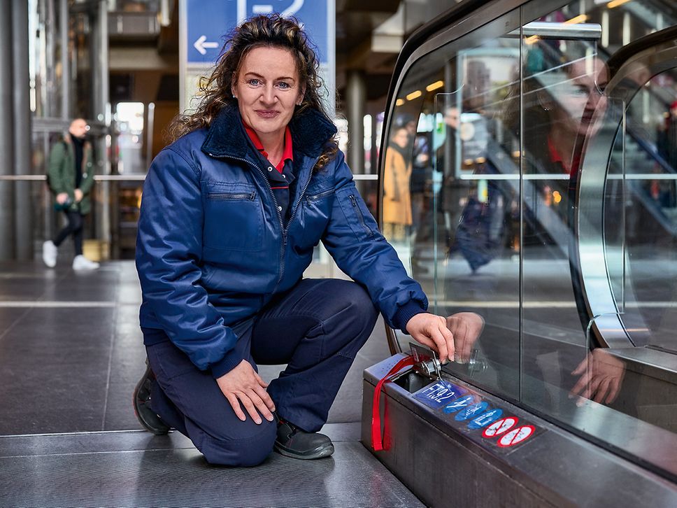 Eine Servicetechnikerin in Arbeitsbekleidung kniet am Bahnhof neben einer Rolltreppe