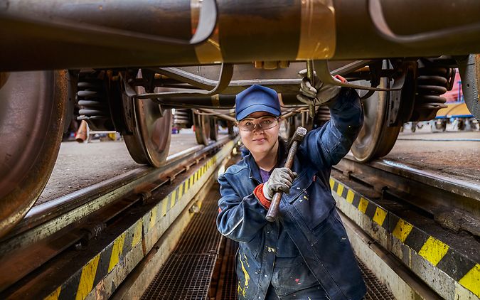 Eine Industriemechanikerin in Arbeitskleidung steht in der Wartungsgrube unter einem Zug und hält einen Hammer