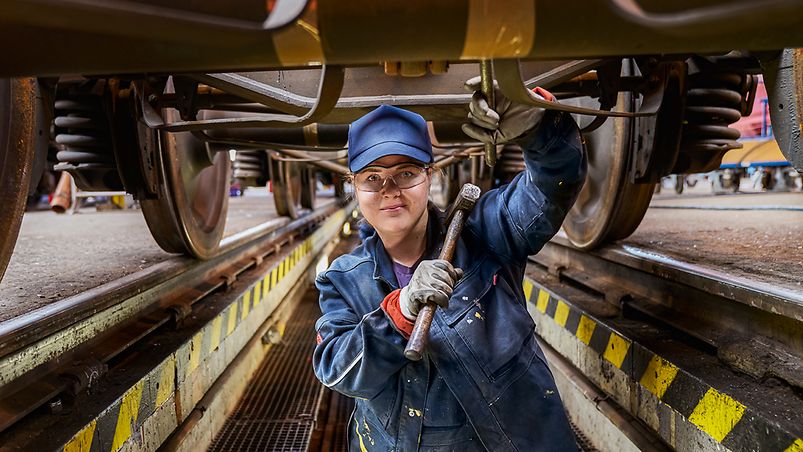 Eine Industriemechanikerin in Arbeitskleidung steht in der Wartungsgrube unter einem Zug und hält einen Hammer