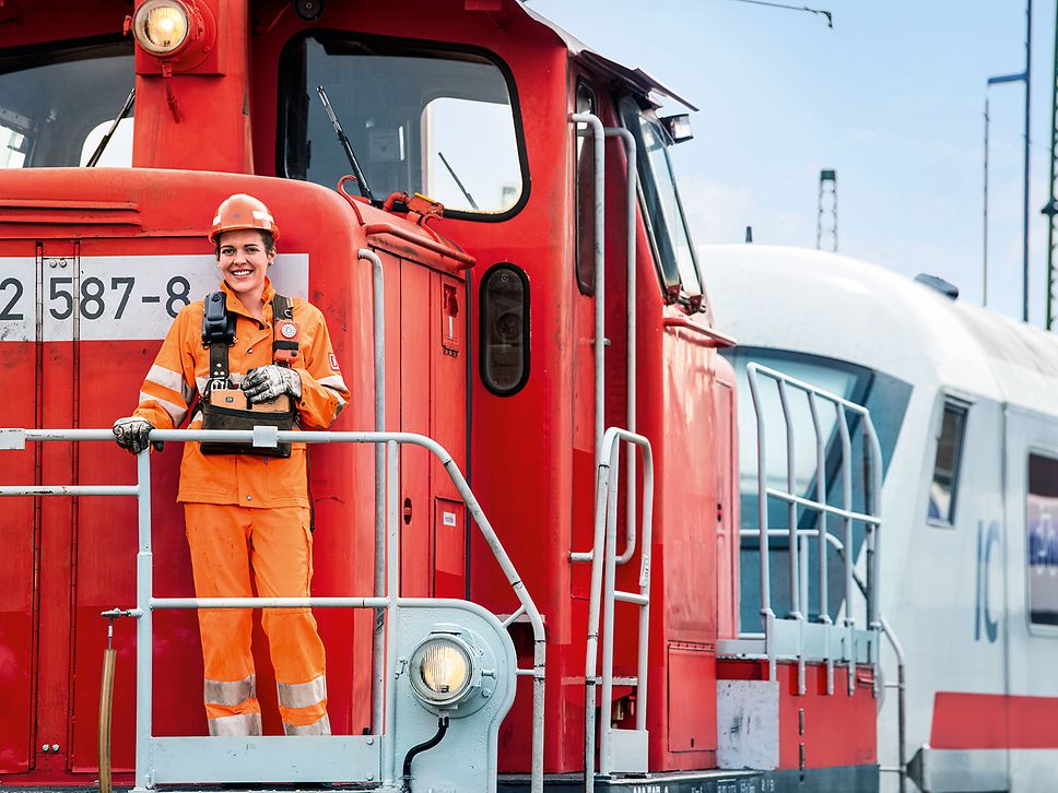 Eine Lokrangierführerin steht in Warnkleidung und mit Schutzhelm auf dem Außenpult des Führerstandes eines Triebwagens