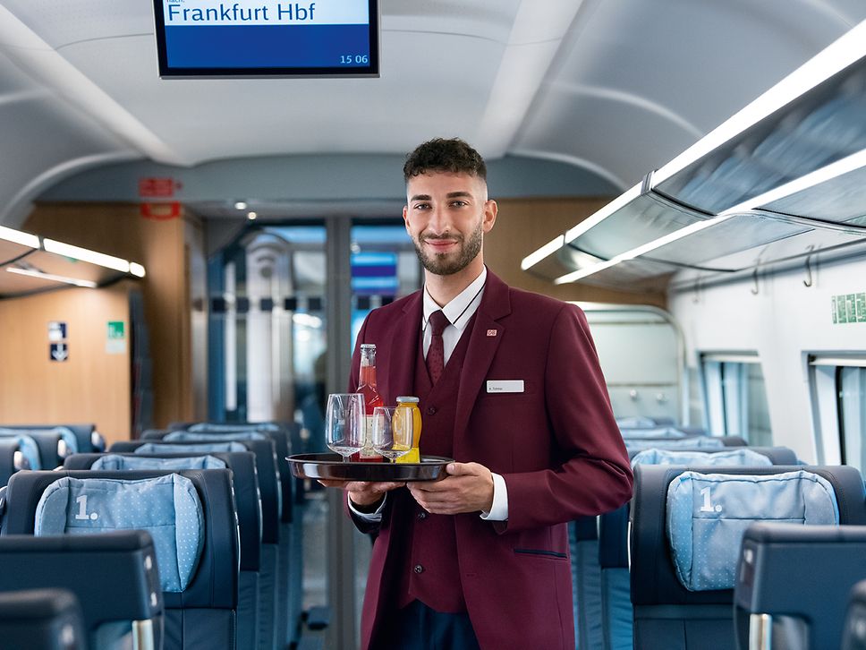 Ein Steward in Unternehmensbekleidung steht mit einem Tablett mit Getränken im Gang eines Zuges