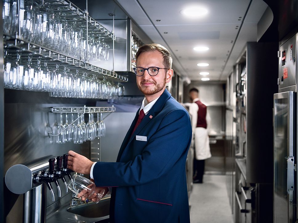 Ein Steward in Unternehmensbekleidung steht im Bordbistro und zapft ein Getränk