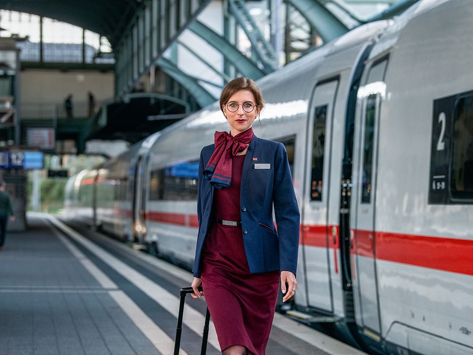 Eine Zugbegleiterin in Unternehmensbekleidung steht am Gleis neben einem ICE
