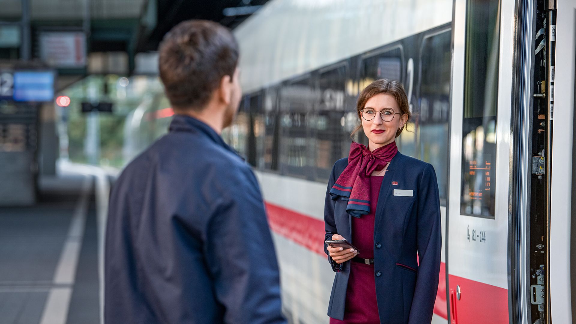 Eine Zugbegleiterin in Unternehmensbekleidung steht am Gleis neben einem ICE und gibt einem Reisenden Auskunft