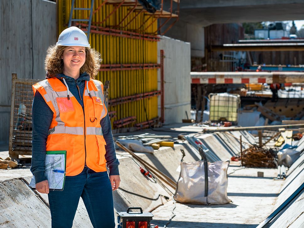 Eine Projektingenieurin steht in Warnkleidung und mit Schutzhelm vor einer Baustelle