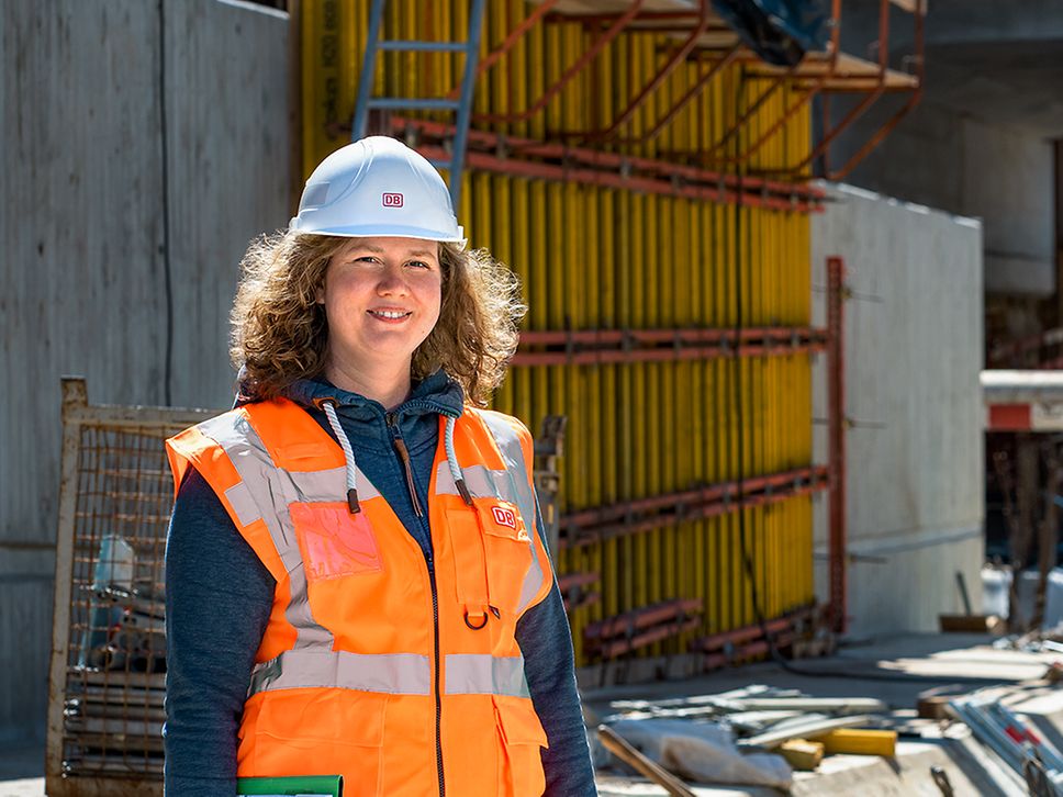 Eine Projektingenieurin steht in Warnkleidung und mit Schutzhelm vor einer Baustelle