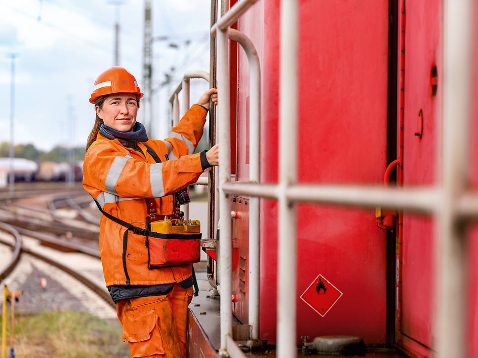 Eine Lokrangierführerin in Warnkleidung und mit Schutzhelm steht auf dem Außenpult eines Güterzuges