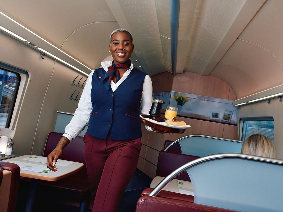 Eine Bordrestaurantleiterin in Unternehmensbekleidung läuft durch den Gang des Bordbistros und trägt ein Tablett mit Getränken