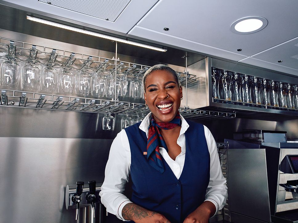 Eine Bordrestaurantleiterin in Unternehmensbekleidung steht im Bordbistro und lächelt herzlich