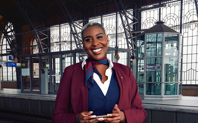 Eine Bordrestaurantleiterin steht an einem Bahnhofsgleis und lächelt.