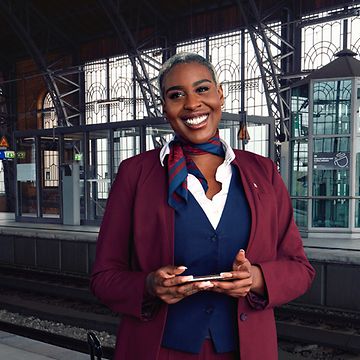 Eine Bordrestaurantleiterin steht an einem Bahnhofsgleis und lächelt.
